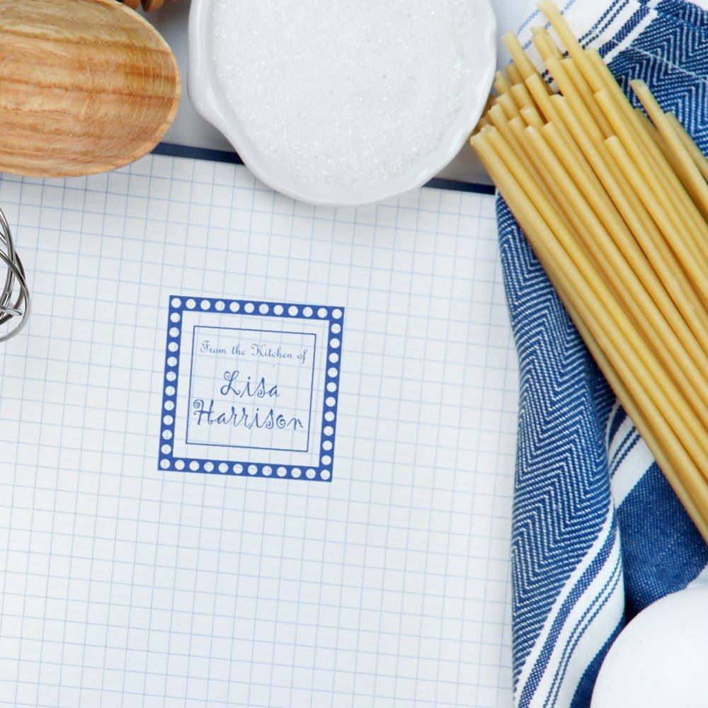 Personalized rubber stamp imprint with name and border on recipe paper, styled with kitchen utensils for custom kitchen branding.