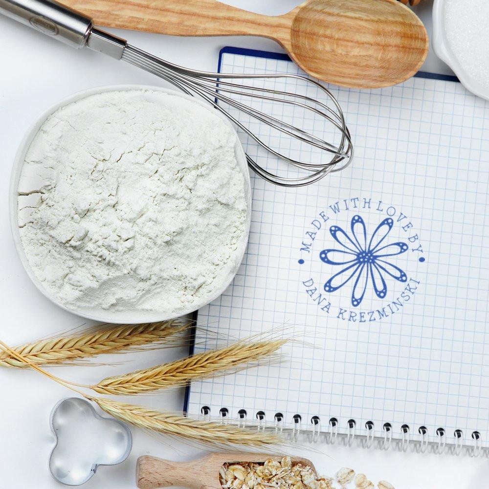 Custom rubber stamp for bakery branding on recipe notebook, surrounded by flour, whisk, and baking tools.