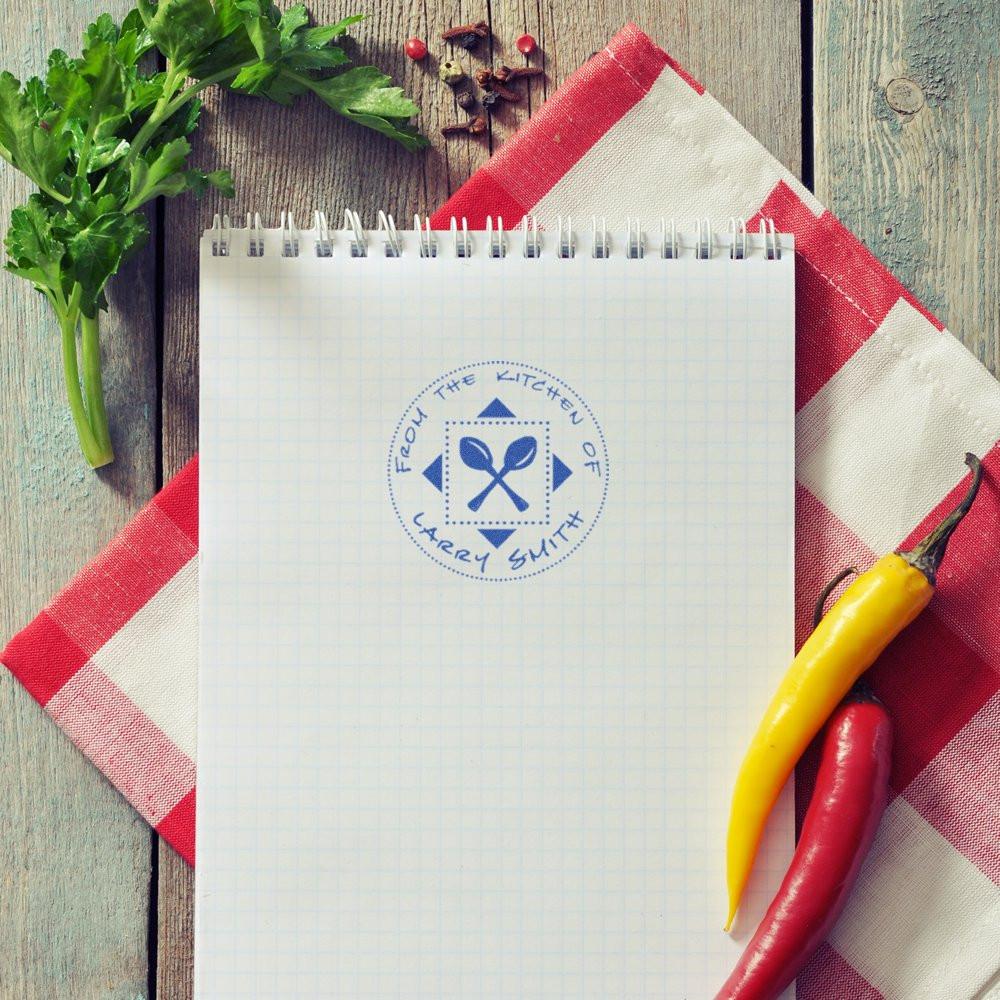 Personalized kitchen rubber stamp on recipe notebook reading From the Kitchen of Larry Smith with crossed spoons, herbs and peppers.