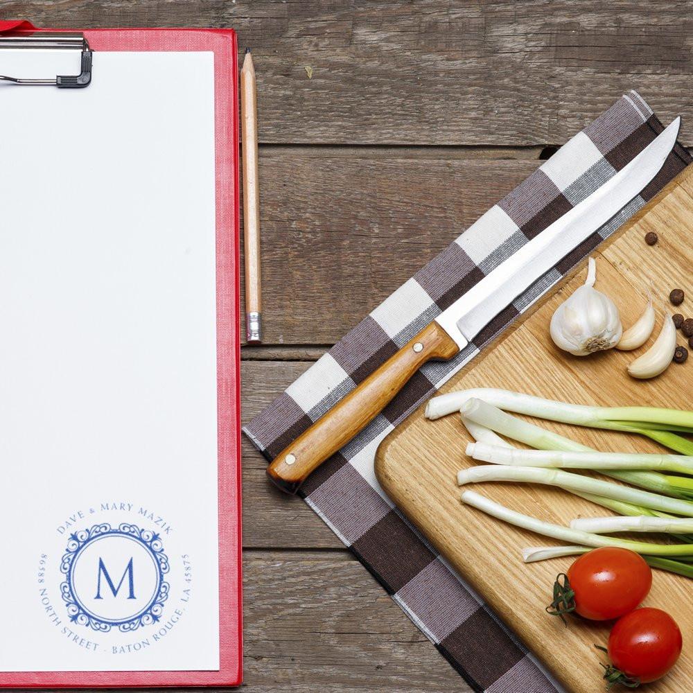 Custom monogram rubber stamp on clipboard beside kitchen ingredients, ideal for personalized recipe cards and stationery branding.