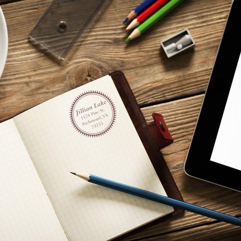 Custom return address rubber stamp on open notebook, with pencils and tablet, for personalized stationery and mail branding.