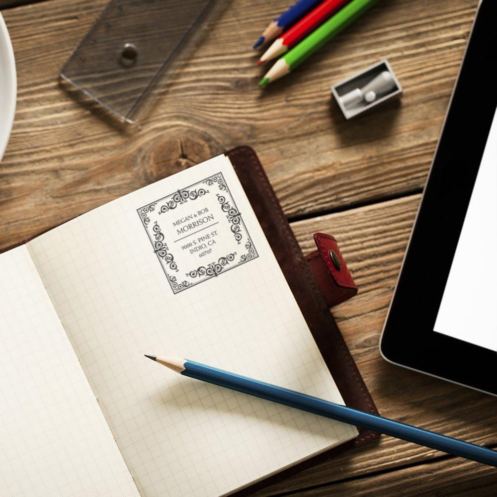 Custom return address rubber stamp imprint on a notebook page beside pencils and tablet on a rustic wooden desk.