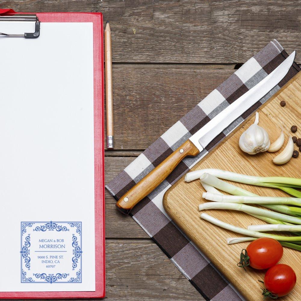 Custom address rubber stamp on clipboard beside kitchen prep board, showing personalized return address stamp design.
