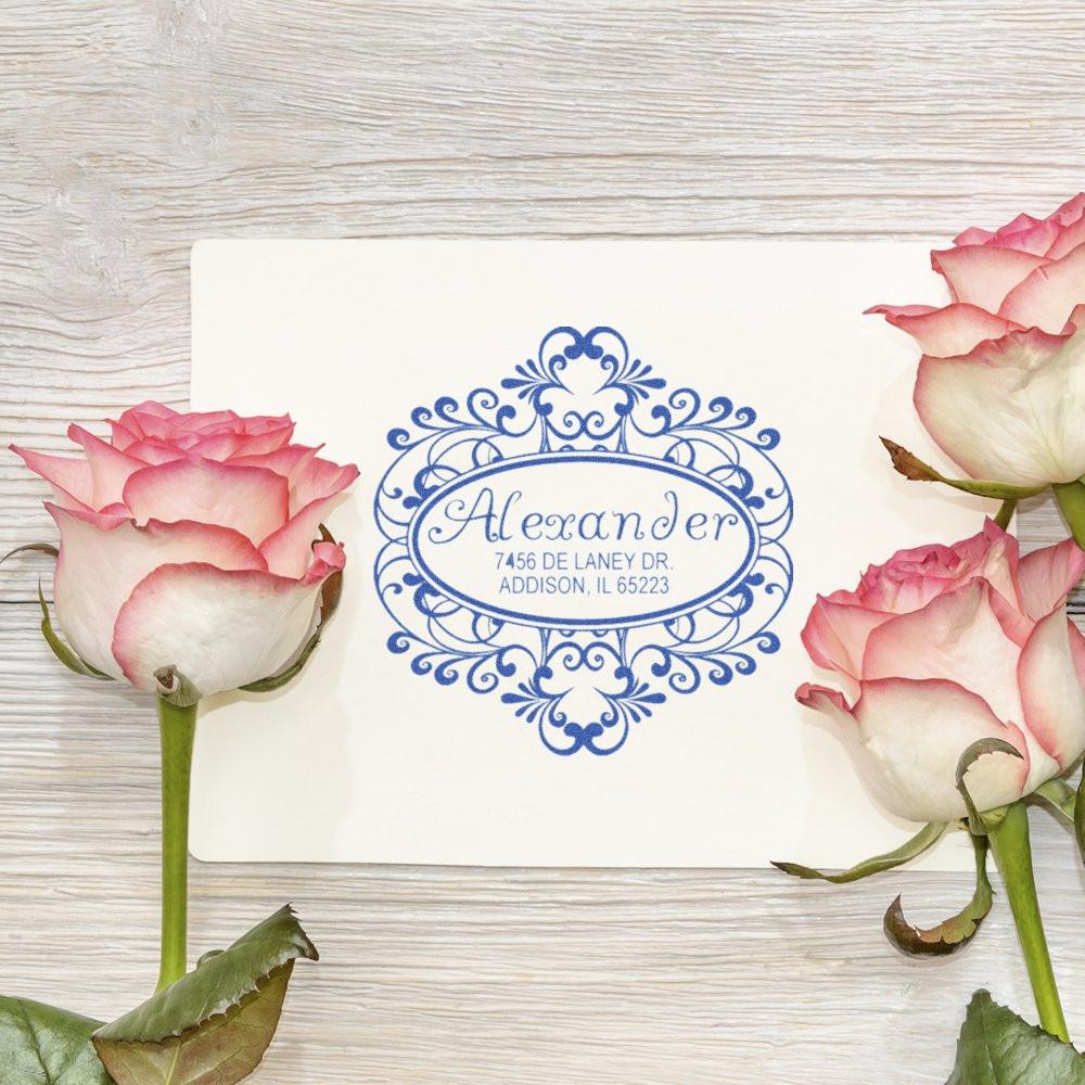Personalized return address rubber stamp with ornate blue floral monogram and name, styled with pink roses on a rustic wood background.