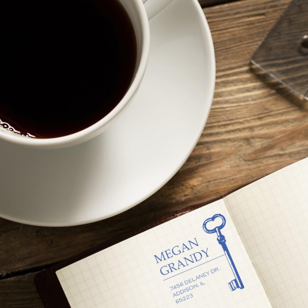 Custom address rubber stamp with key design on notebook beside coffee cup, ideal for personalized stationery and mail.
