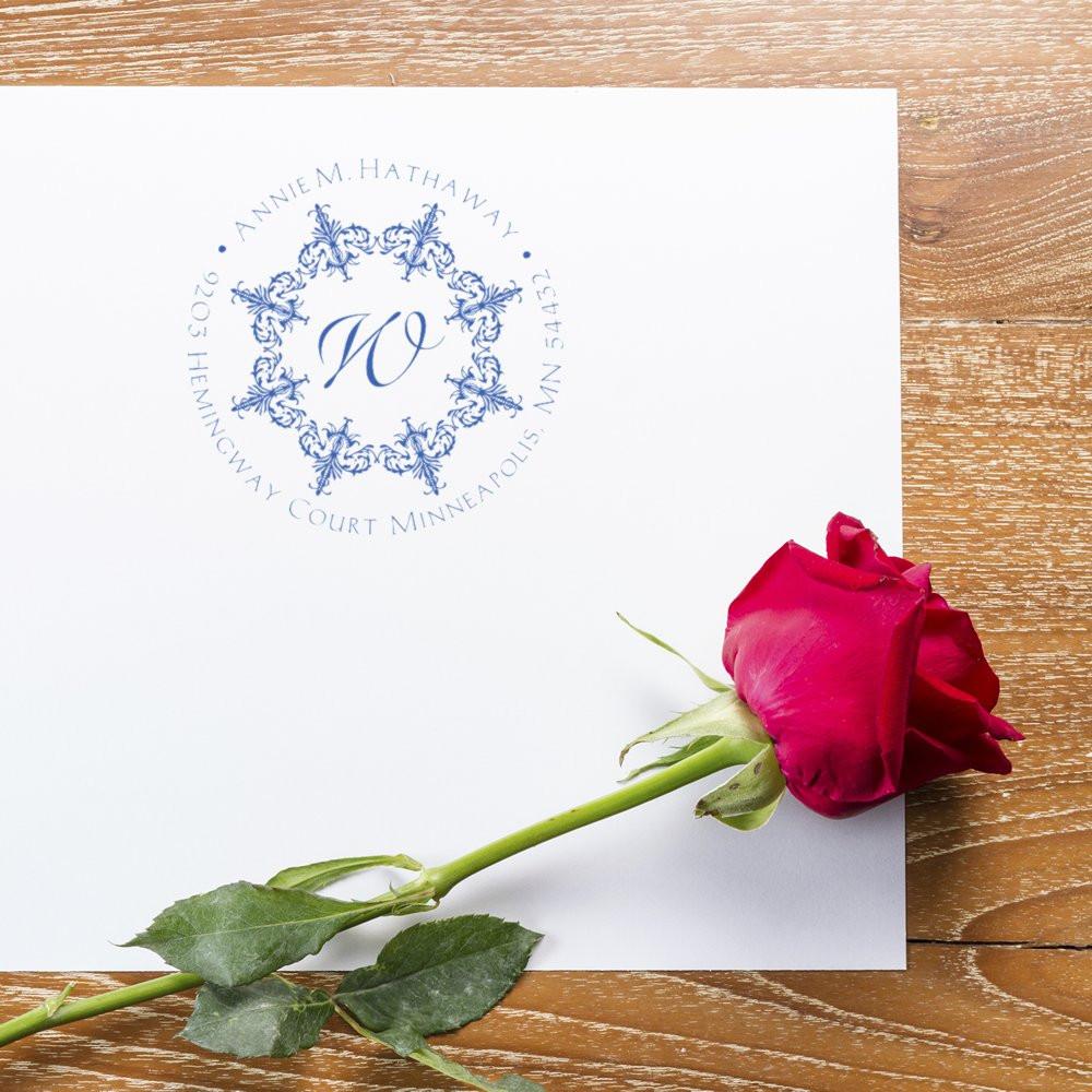 Personalized return address rubber stamp with floral monogram, name, and address on wedding stationery beside a red rose.