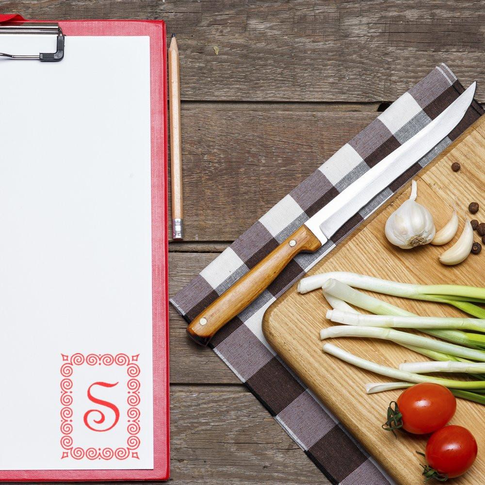 Recipe book beside chef knife with plaid cloth, garlic, tomatoes and scallions on wooden cutting board for fresh meal prep kitchen.