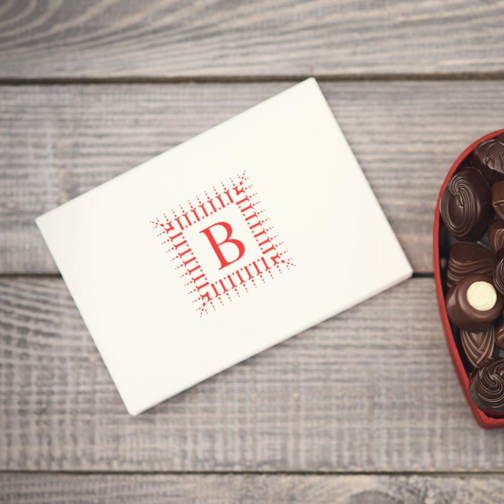Custom monogram rubber stamp with red letter B on white envelope, beside chocolates on a rustic wood table.
