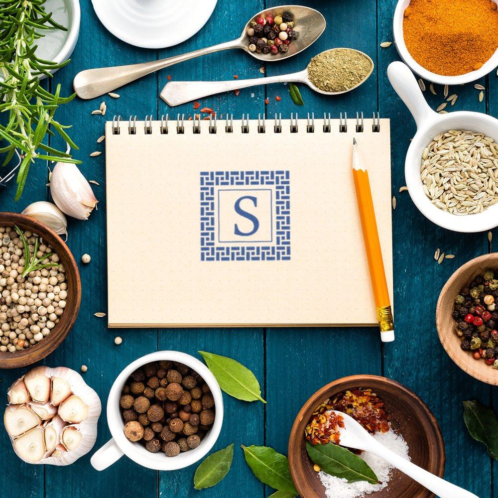 Custom monogram rubber stamp with letter S on a recipe notebook, styled with spices and herbs for branded kitchen labeling.