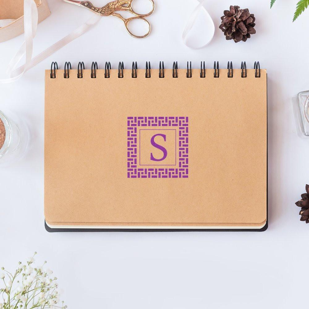 Custom monogram rubber stamp with purple initial S on kraft notebook for personalized stationery and branding.