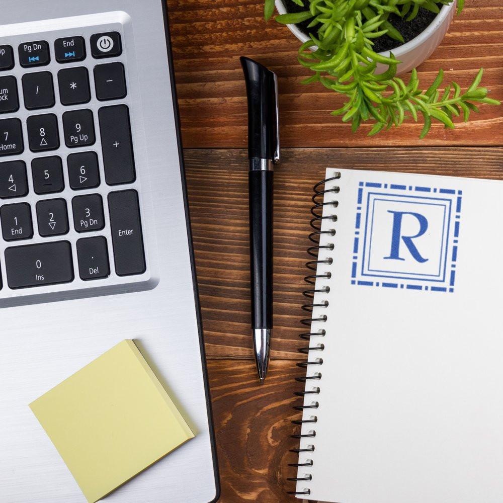 Blue custom rubber stamp impression with letter R on notebook, on office desk beside laptop and pen.