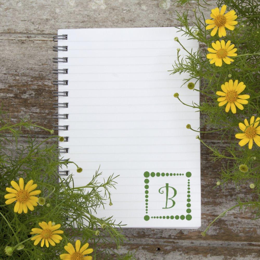 Blank spiral notebook with floral monogram B on rustic wood, framed by yellow daisies, ideal for custom stationery.
