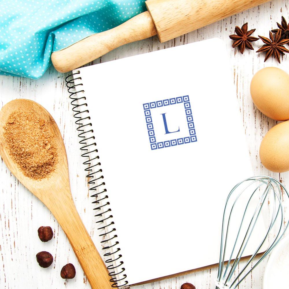 Custom monogram rubber stamp with letter L on a recipe notebook, surrounded by baking tools for personalized kitchen branding.
