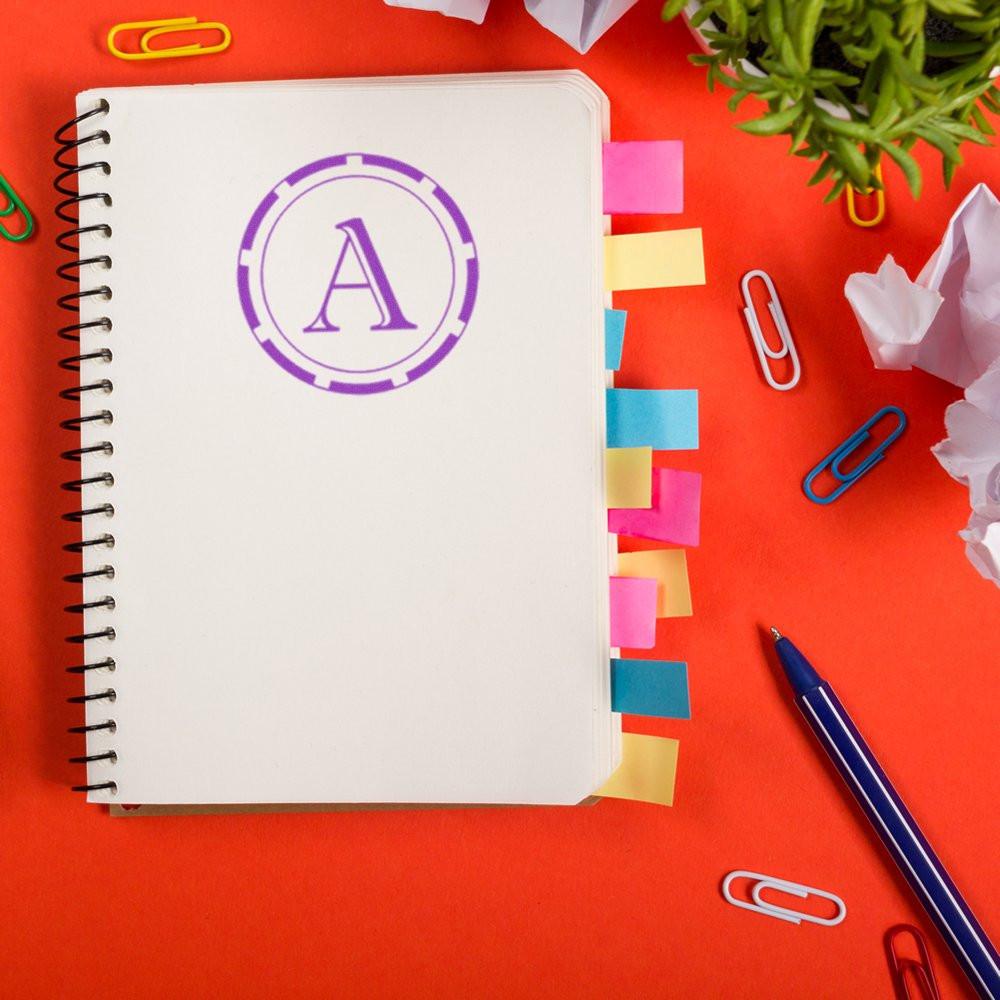 Custom monogram rubber stamp with letter A on a notebook page, shown as a branding sample with office supplies.