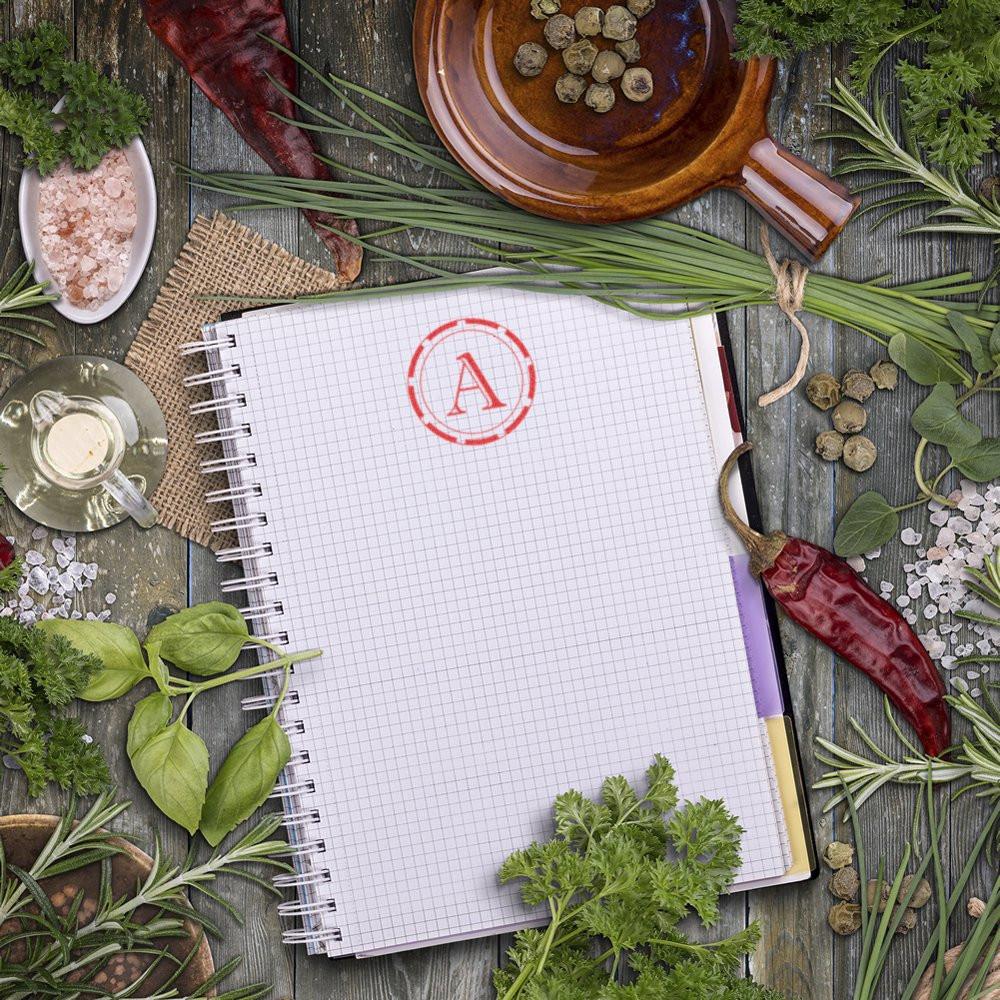 Monogram rubber stamp with red letter A imprint on notebook, surrounded by herbs and spices on rustic wood.