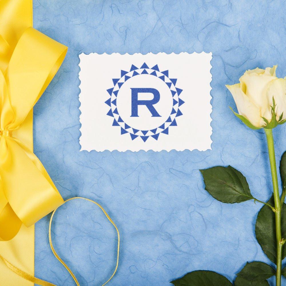 Custom rubber stamp with blue monogram R logo on scalloped white paper, styled with yellow ribbon and white rose.
