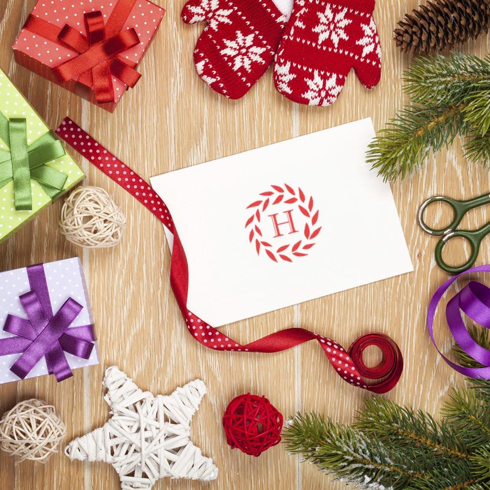 Custom holiday rubber stamp with monogram H wreath on card, surrounded by gift wrap, ribbon, and festive Christmas decor.