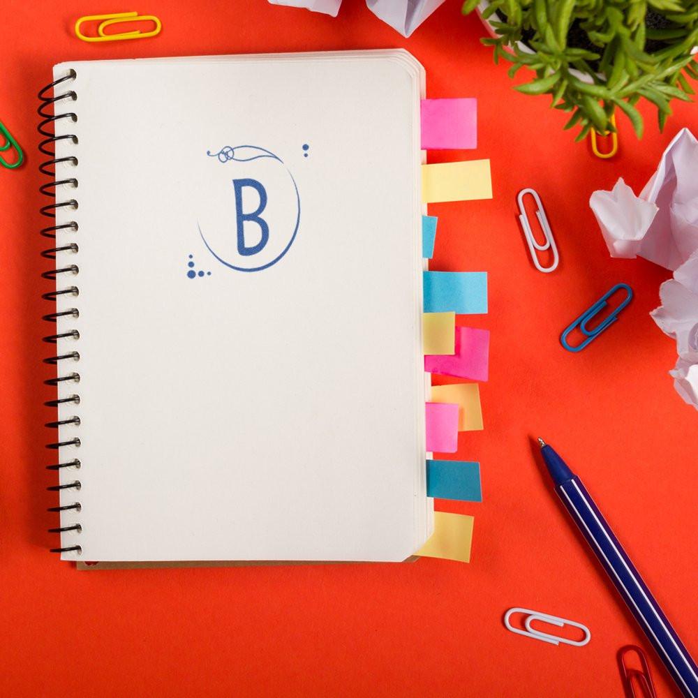 Custom monogram B rubber stamp on spiral notebook, surrounded by colorful tabs and office supplies.