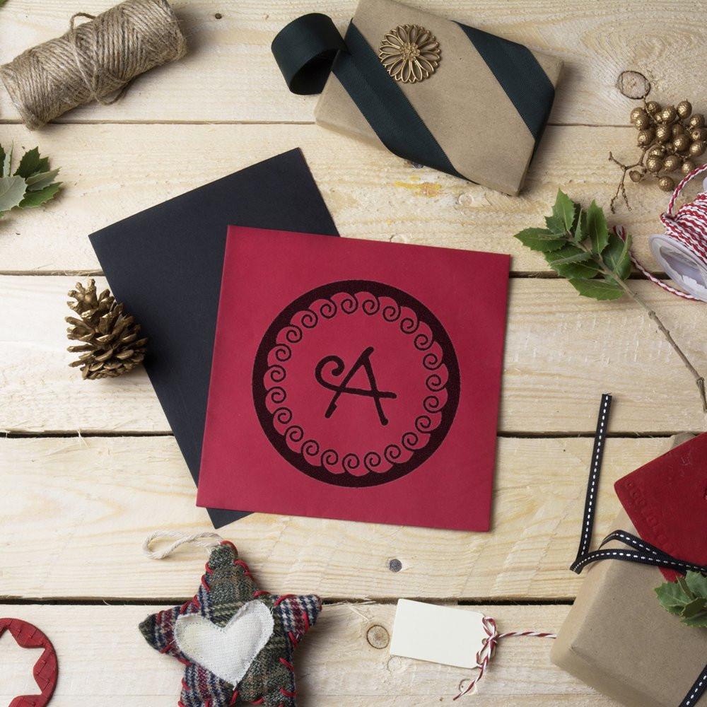 Handmade Christmas greeting card with letter A in red circular monogram, surrounded by wrapped gifts, pine branches, and twine.