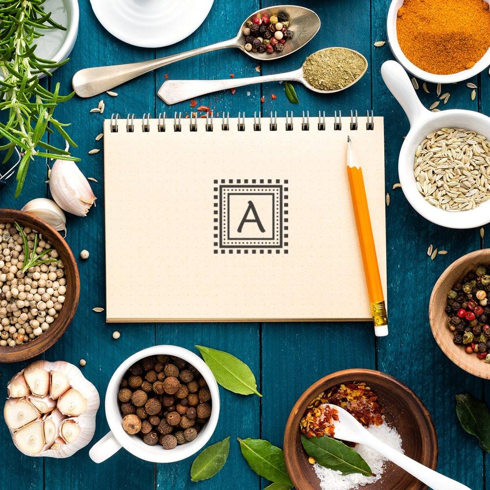Custom alphabet A rubber stamp design on notebook, framed by herbs and spices for culinary craft branding.