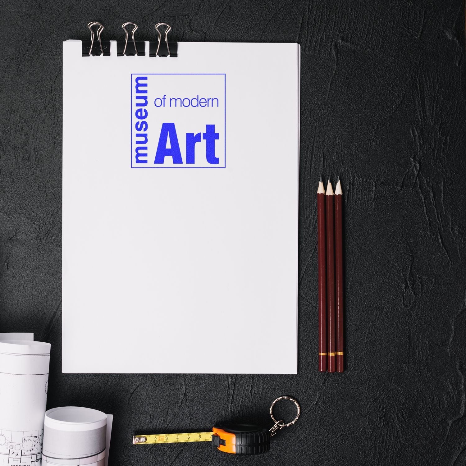 Modern art studio flat lay with Museum of Modern Art sketchbook, artist pencils, tape measure, and keychain on dark background