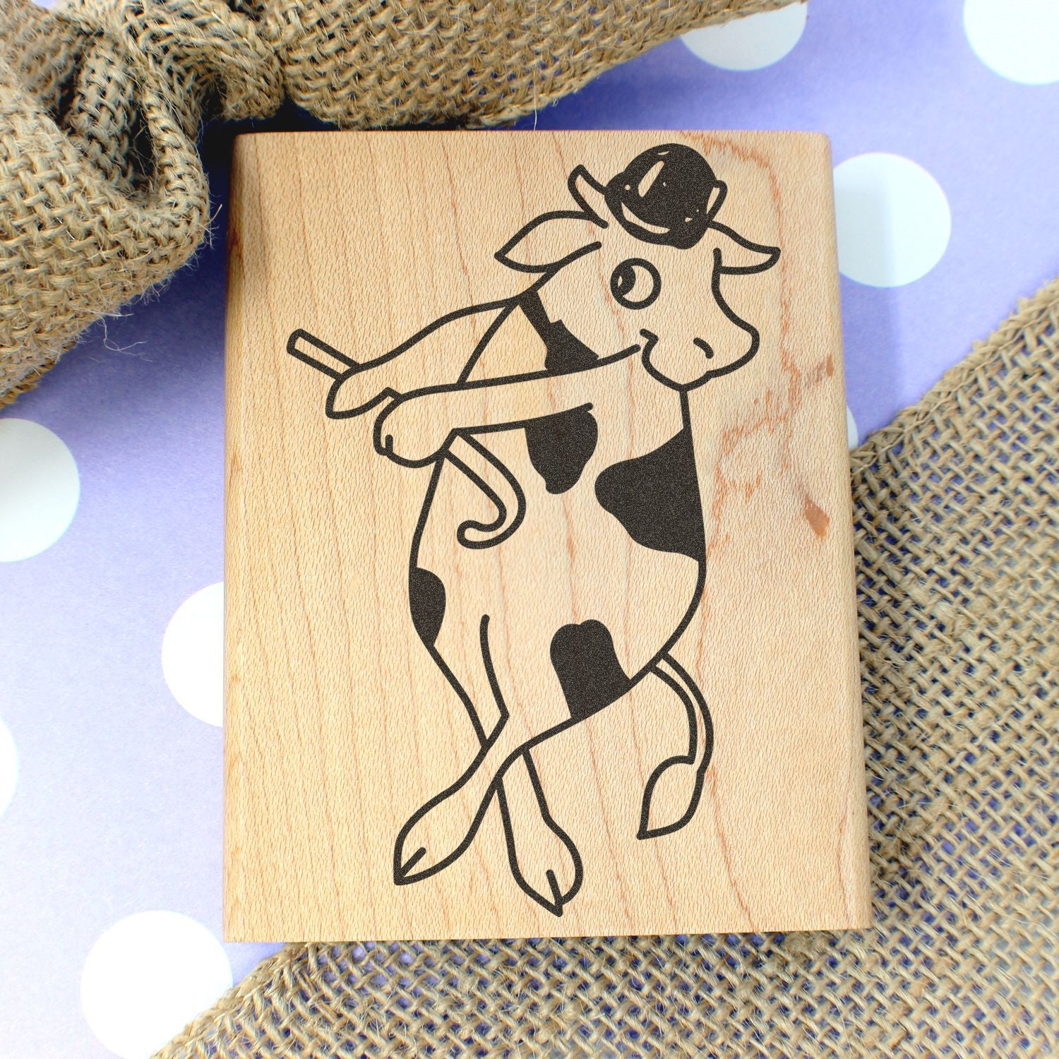 Wooden cow stamp with dancing cow illustration on rustic burlap and purple polka dot background.