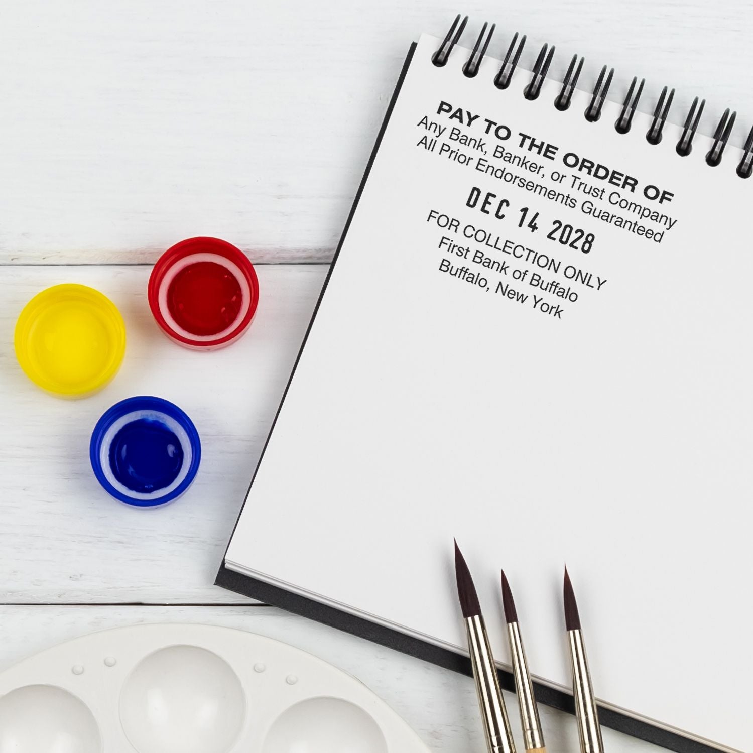 Sketchbook with Pay to the Order of print, watercolor paint pots, brushes, and palette for art journaling and creative painting.