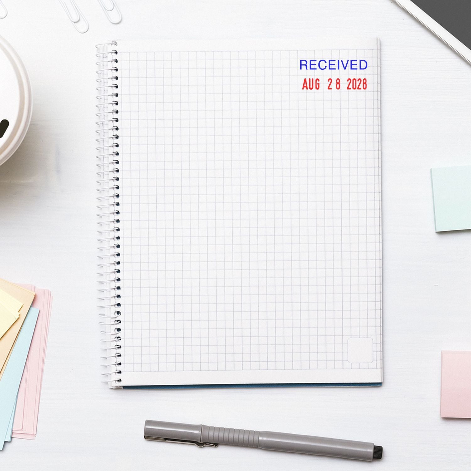 Top view of spiral grid notebook with Received Aug 28 2028 stamp, pen, and colorful sticky notes on a clean white desk.