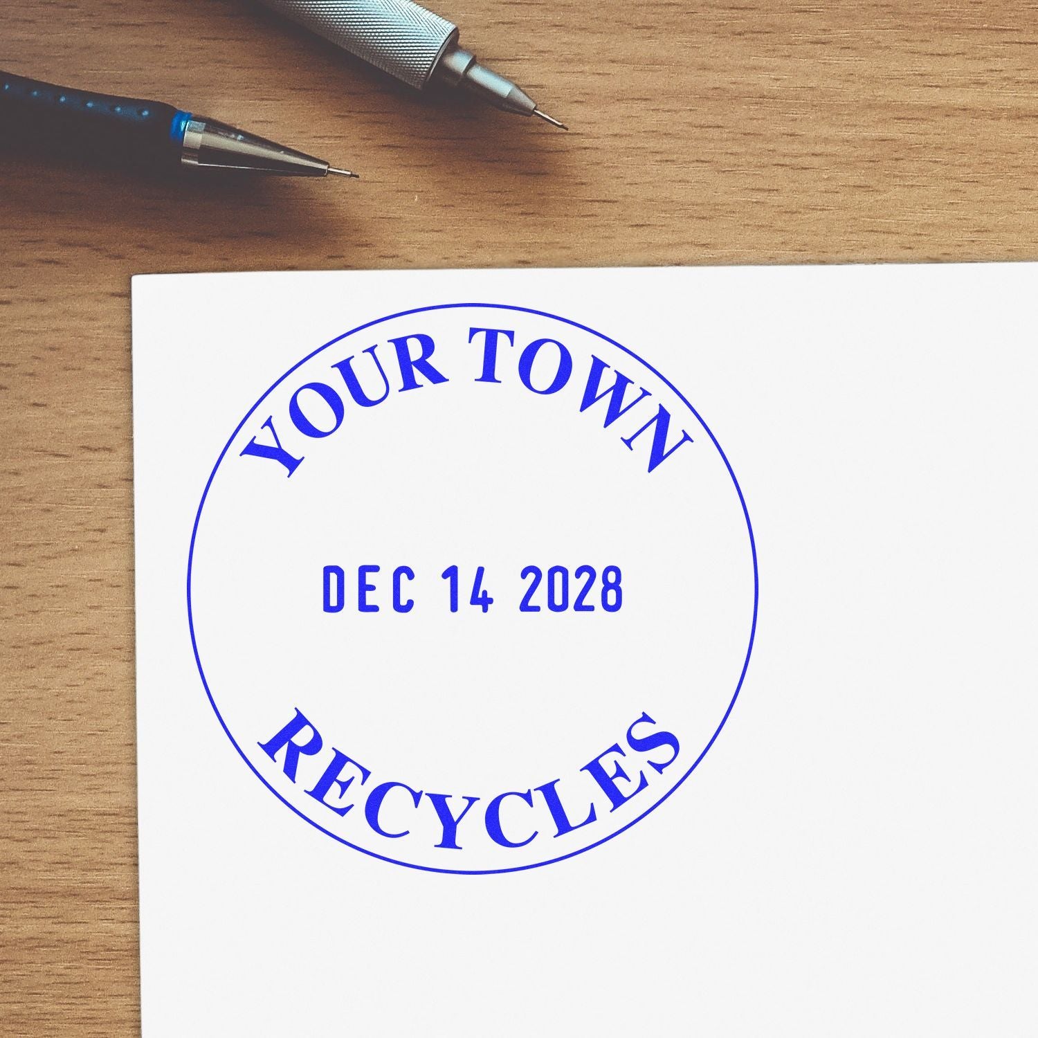 Your Town Recycles date stamp on paper with blue text, pens on wooden desk, and Dec 14 2028 in the center.
