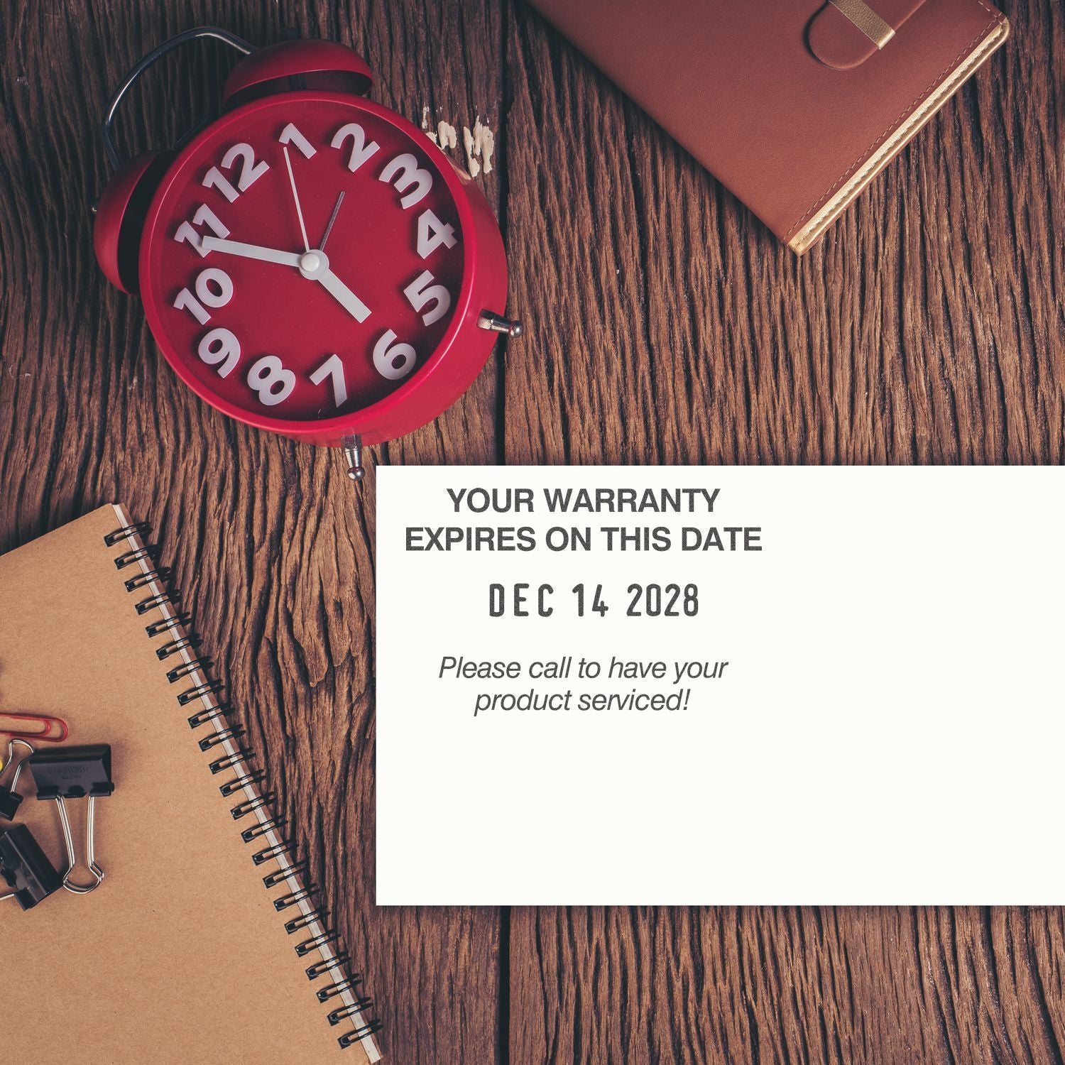 Red alarm clock on wooden desk with notebook and warranty notice reading Your warranty expires on this date, Dec 14 2028 reminder.