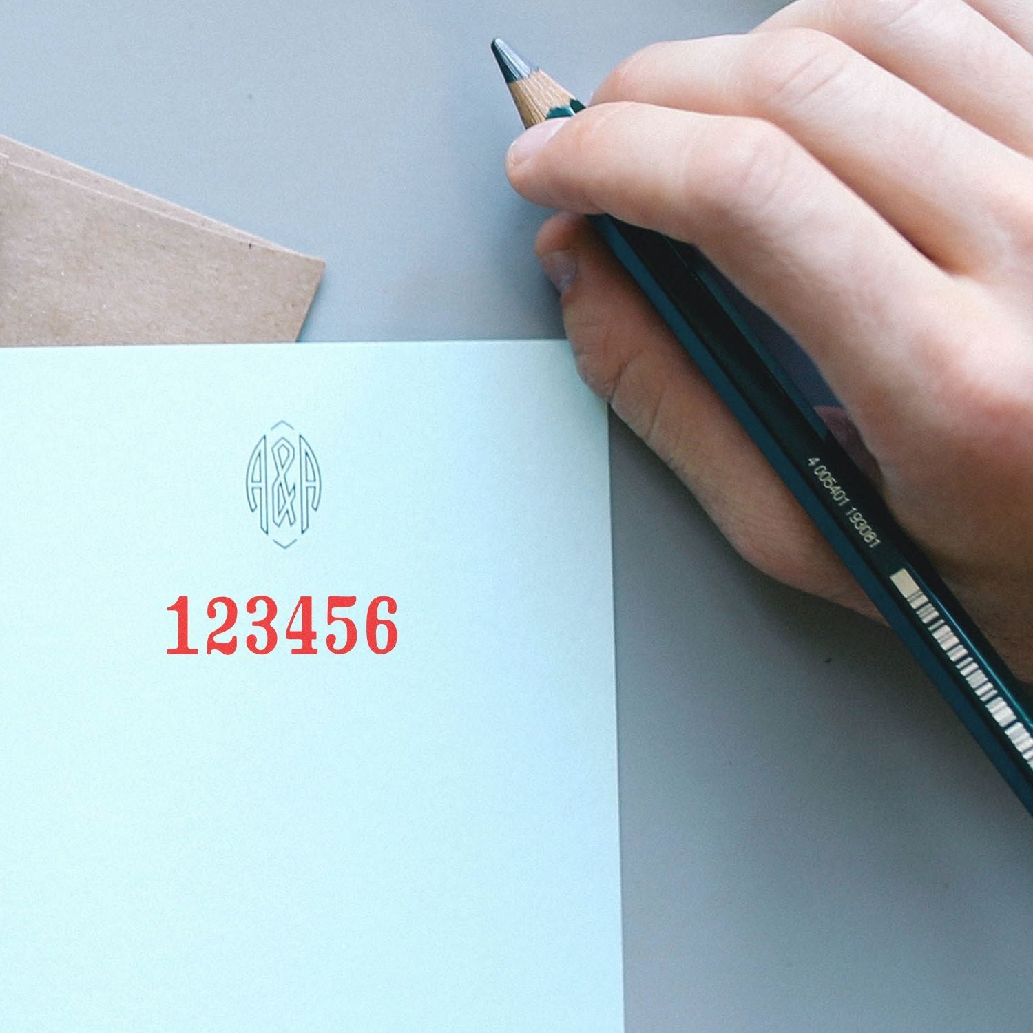 Hand holding a pencil over personalized stationery with monogram logo and red 123456 number design.
