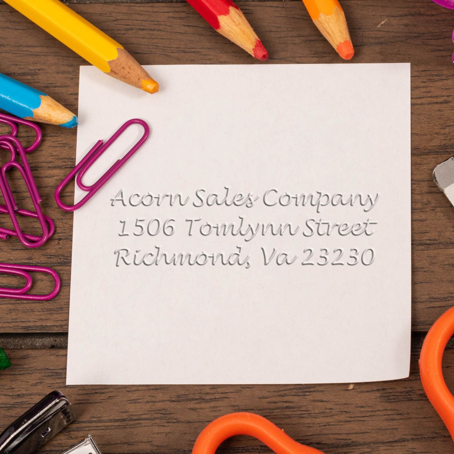 Blank note with Acorn Sales Company address, surrounded by colorful pencils and paper clips on a wooden desk.