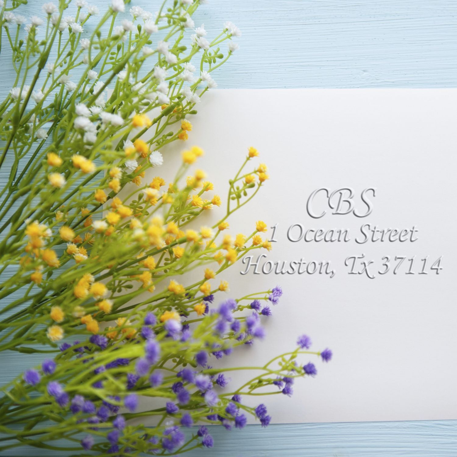 Colorful floral arrangement beside a white embossed invitation card with CBS and the address 1 Ocean Street, Houston, TX 37114 on a light blue wooden background.