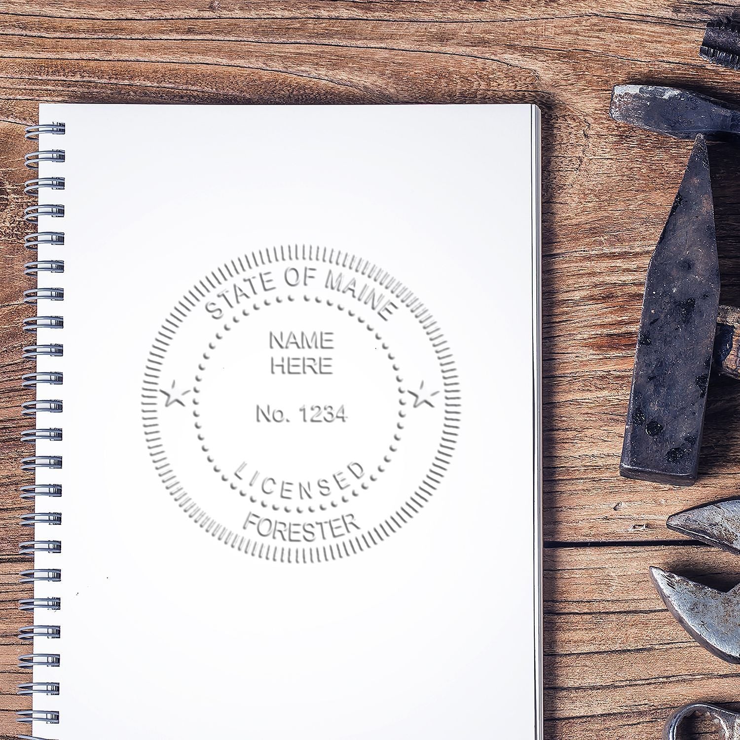 Spiral notebook with State of Maine licensed forester seal on a wooden desk, featuring forestry tools.