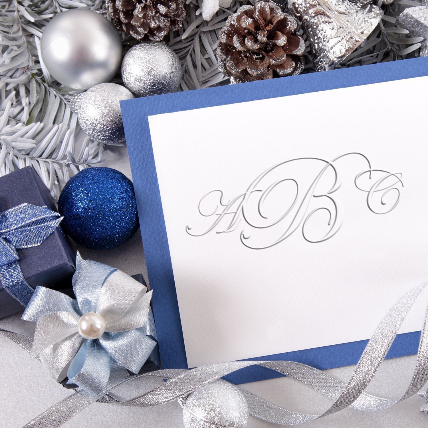 Elegant blue and silver Christmas greeting card with ornaments, pine cones, ribbon, and blank monogrammed card background