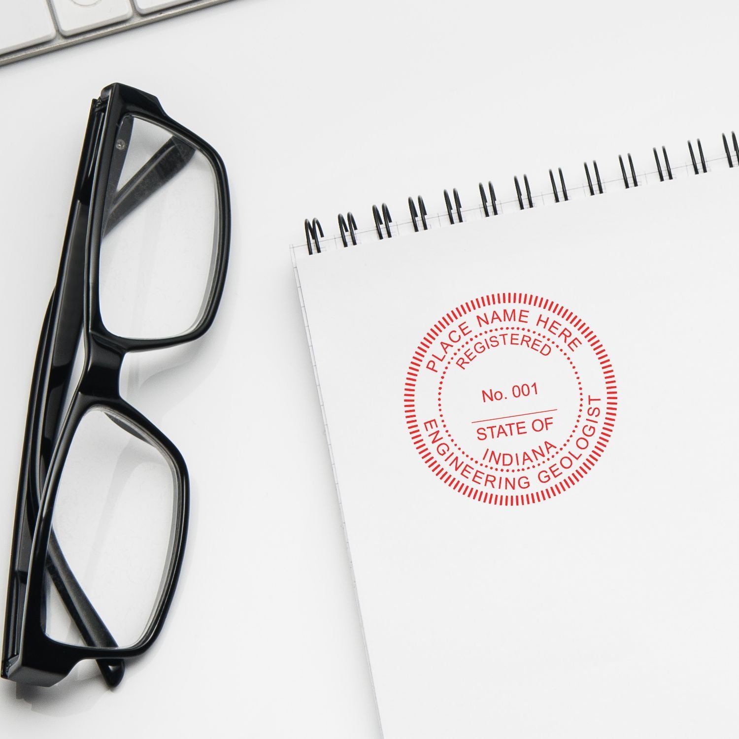 Close-up of eyeglasses beside a spiral notebook with a red engineering geologist seal on a white desk.