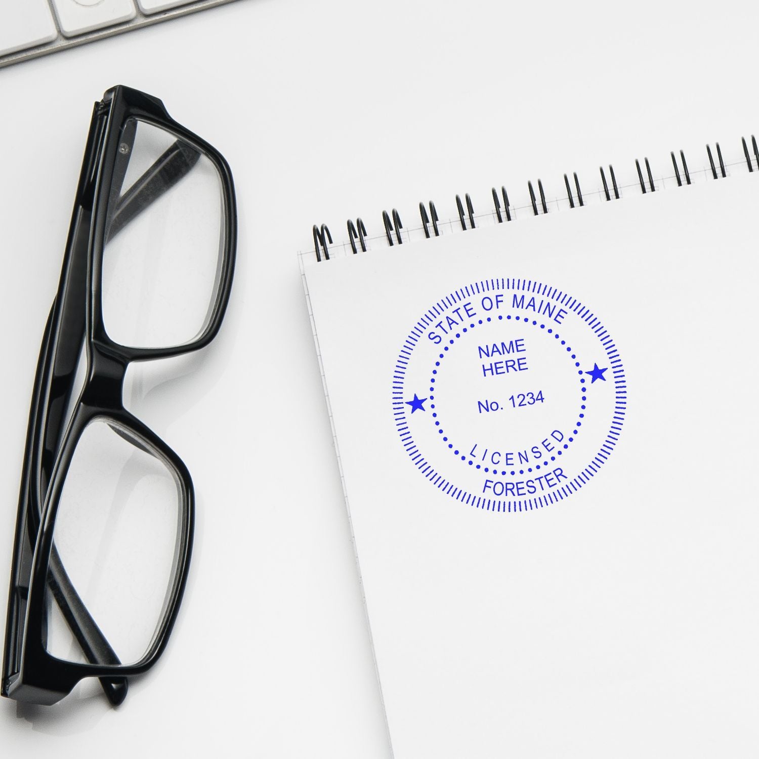 Black eyeglasses beside a spiral notebook with a Maine licensed forester stamp on a white desk.