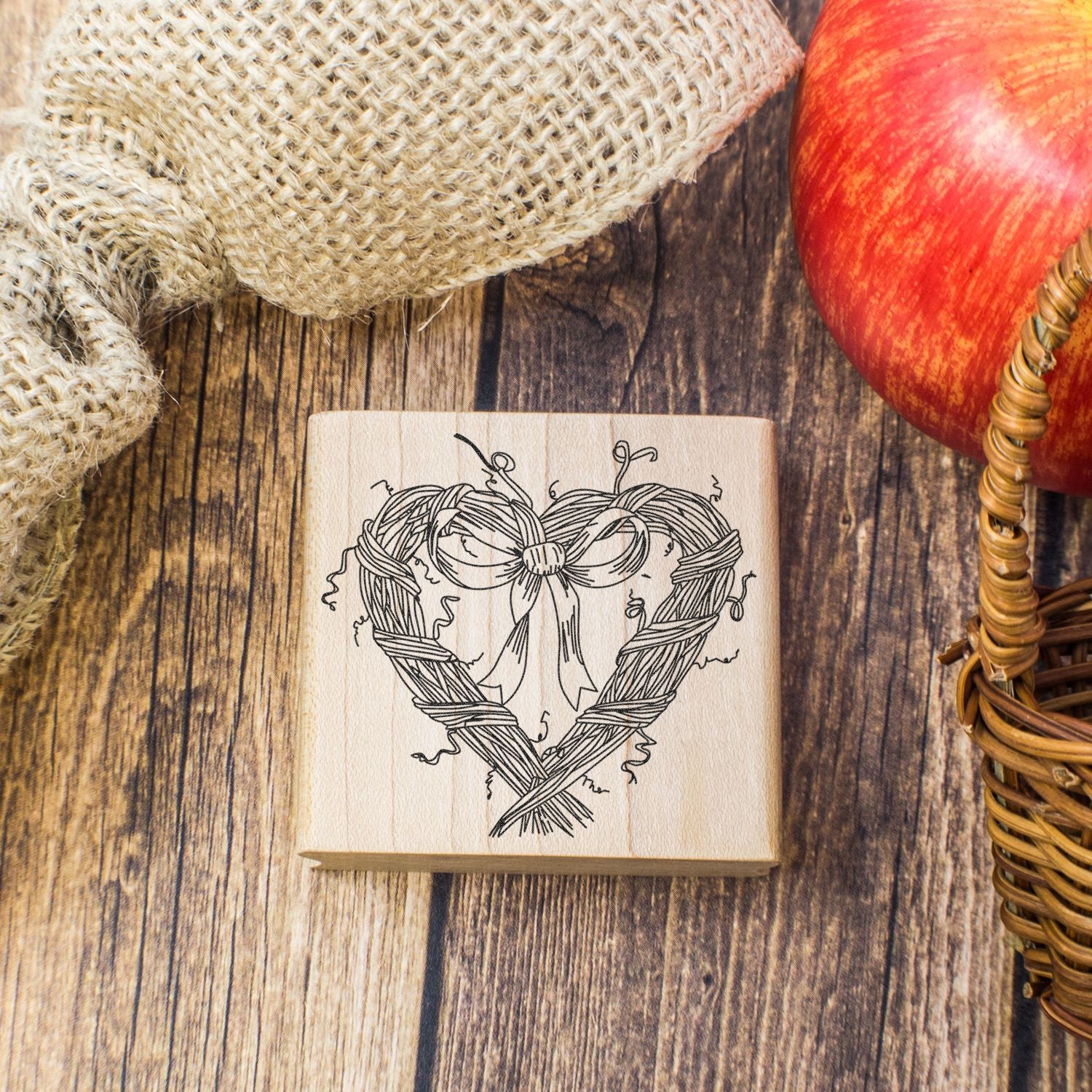 Rustic handmade wooden stamp with woven heart and bow design on a wooden table, styled with burlap and apple decor.