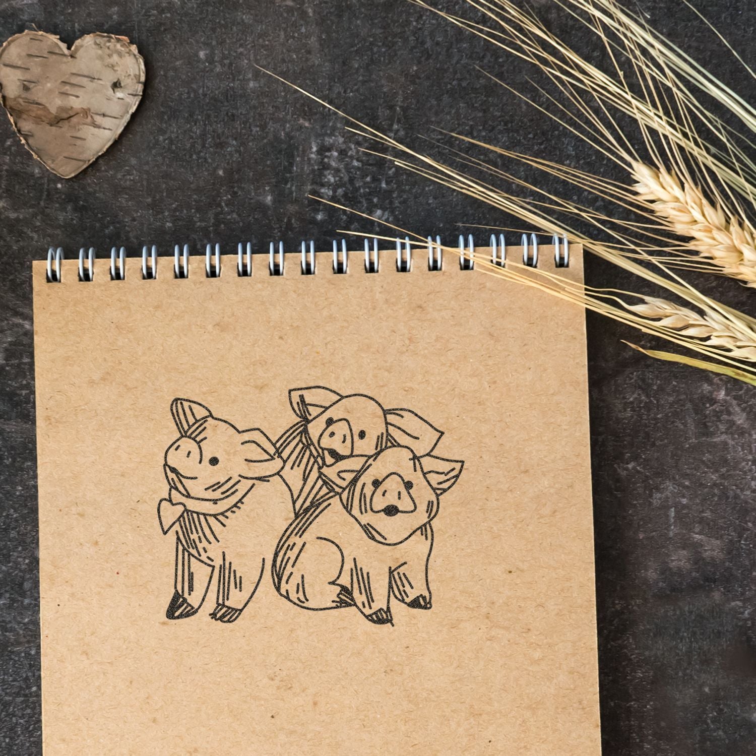 Kraft spiral notebook with cute pig drawing, rustic wheat and birch heart decor on a dark background.