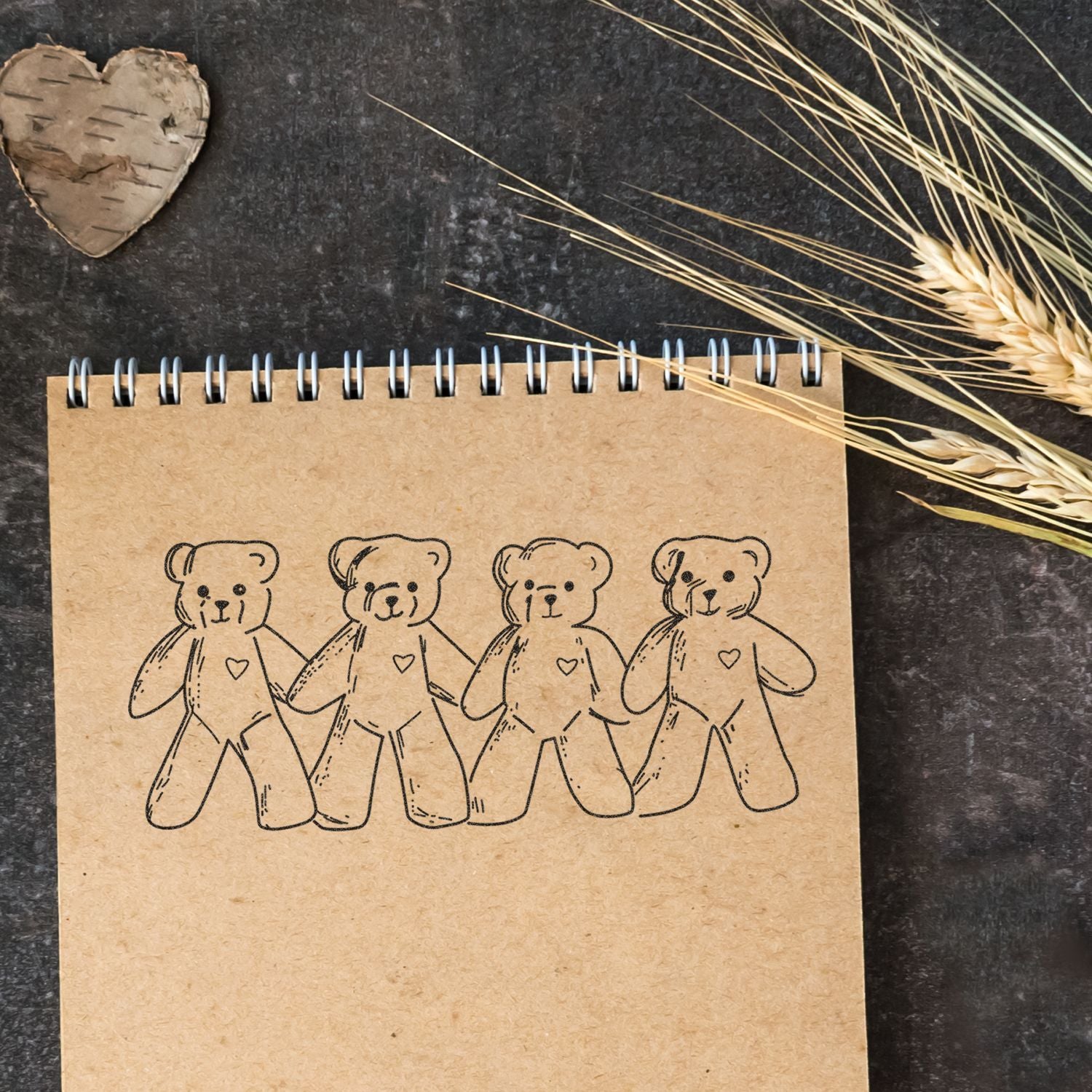 Cute teddy bear illustration on a kraft notebook with dried wheat and rustic heart decor on a dark background.