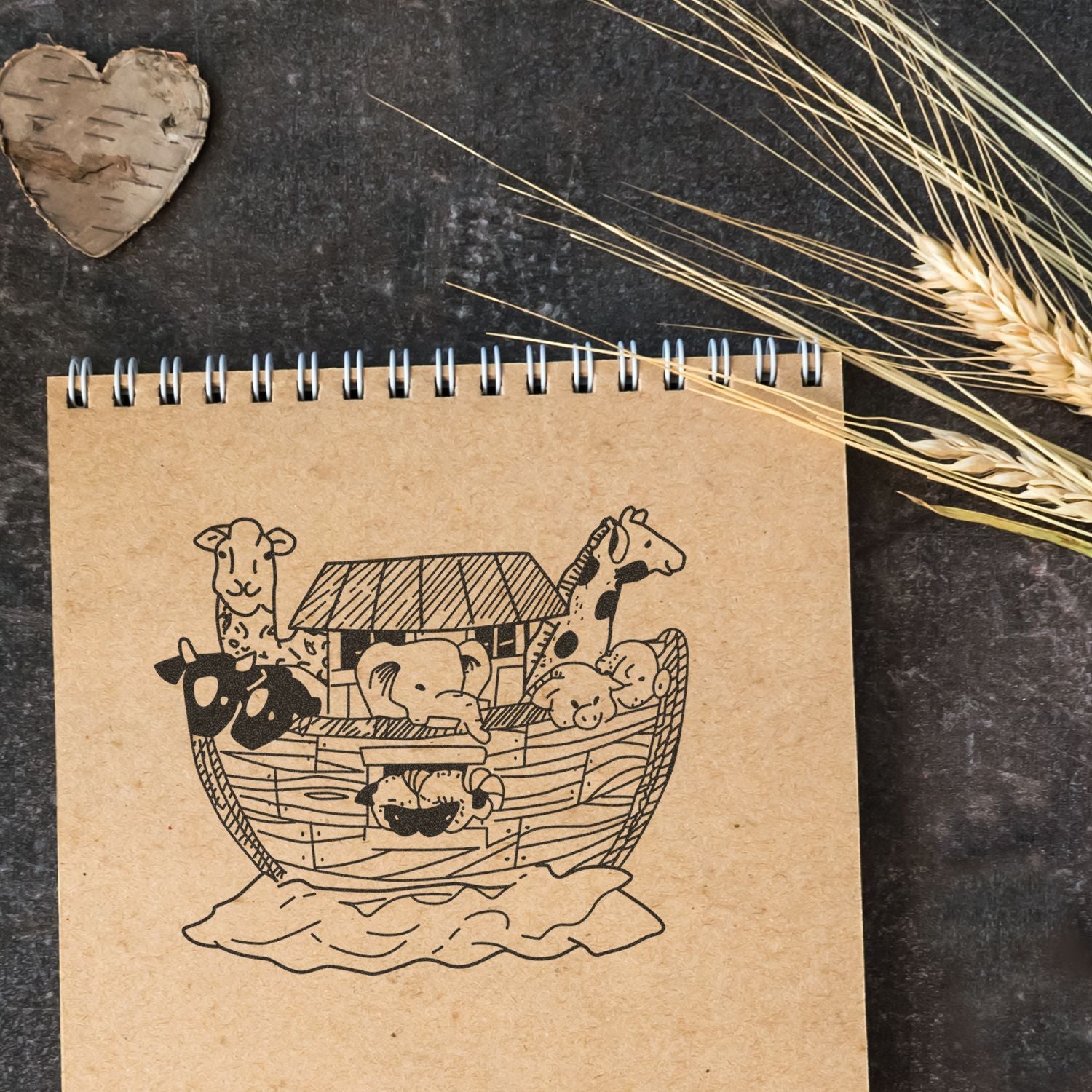 Rustic notebook with Noahs Ark animal illustration, surrounded by wheat stalks and a wooden heart on a dark background.