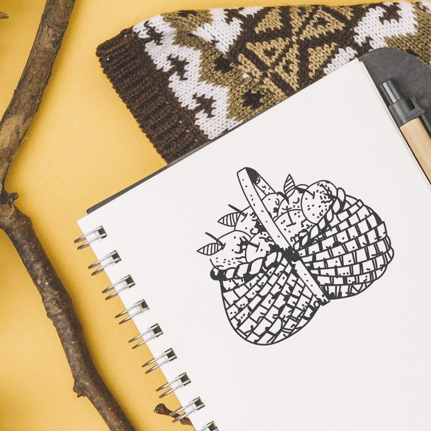 Spiral sketchbook with hand-drawn fruit basket illustration, rustic craft supplies, knitted textile, and pen on a yellow background.