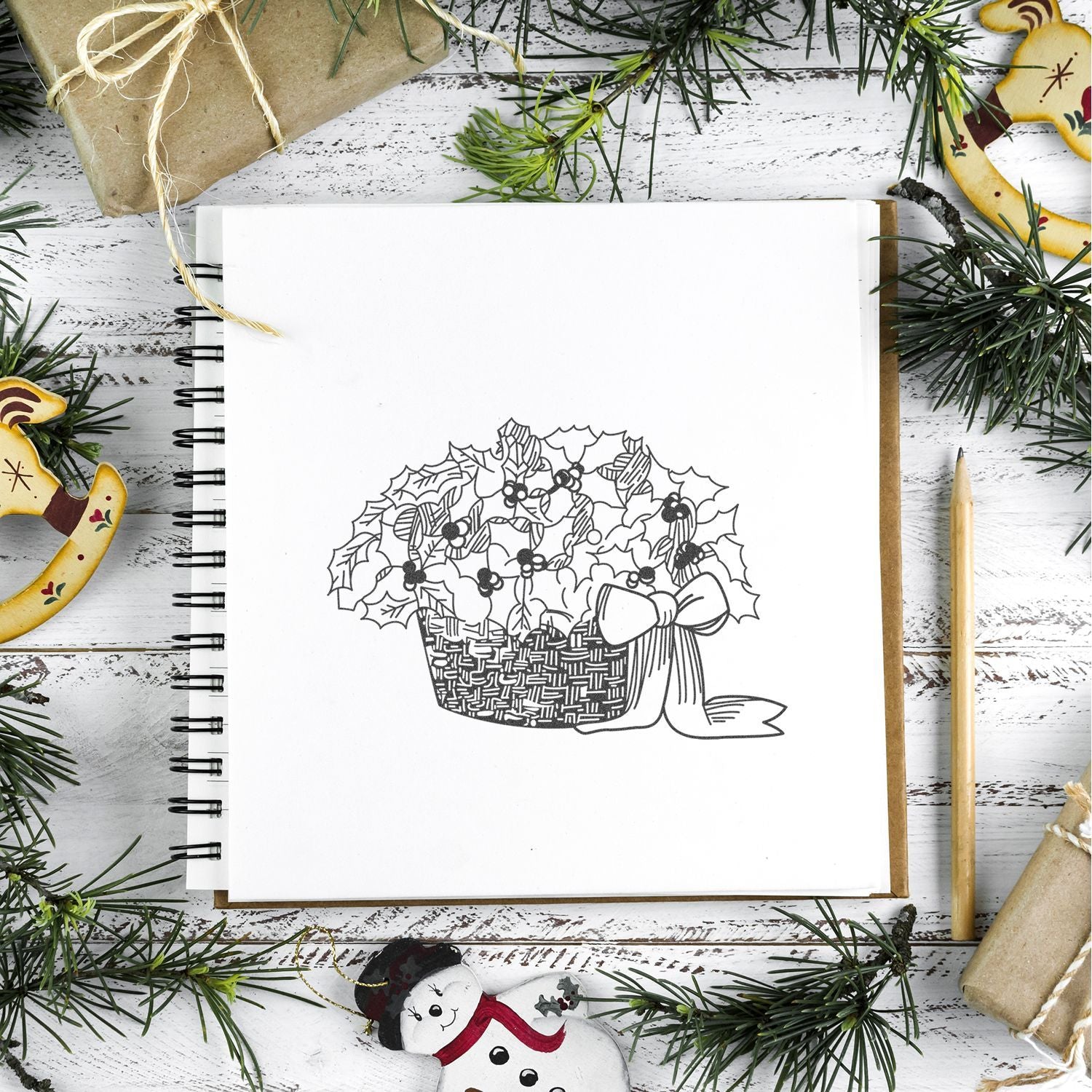 Christmas sketchbook with holly basket drawing, surrounded by evergreen branches, wrapped gifts, and festive holiday decorations on a rustic white wooden background.