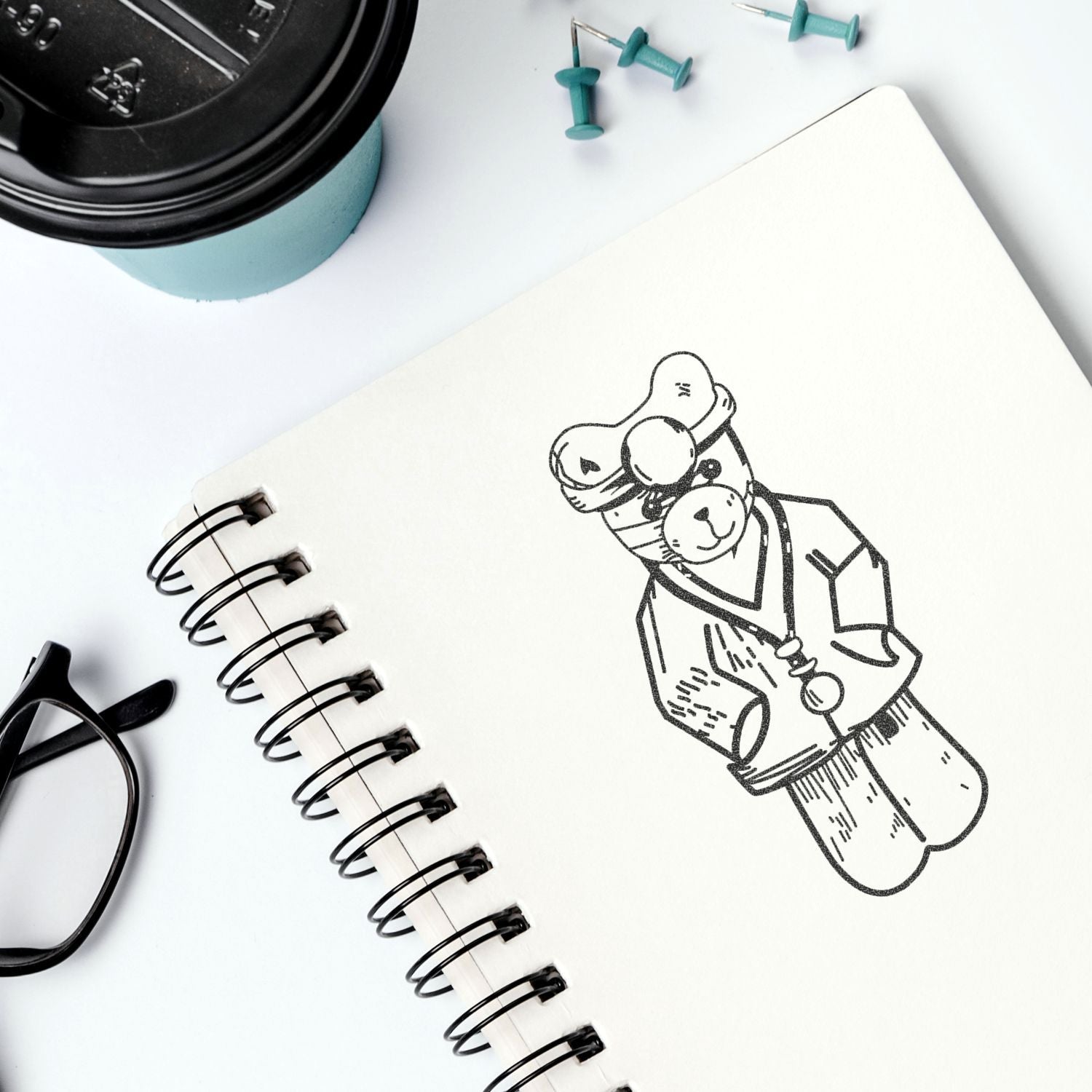 Spiral notebook with cute teddy bear doctor sketch, coffee cup, glasses, and push pins on a white desk.