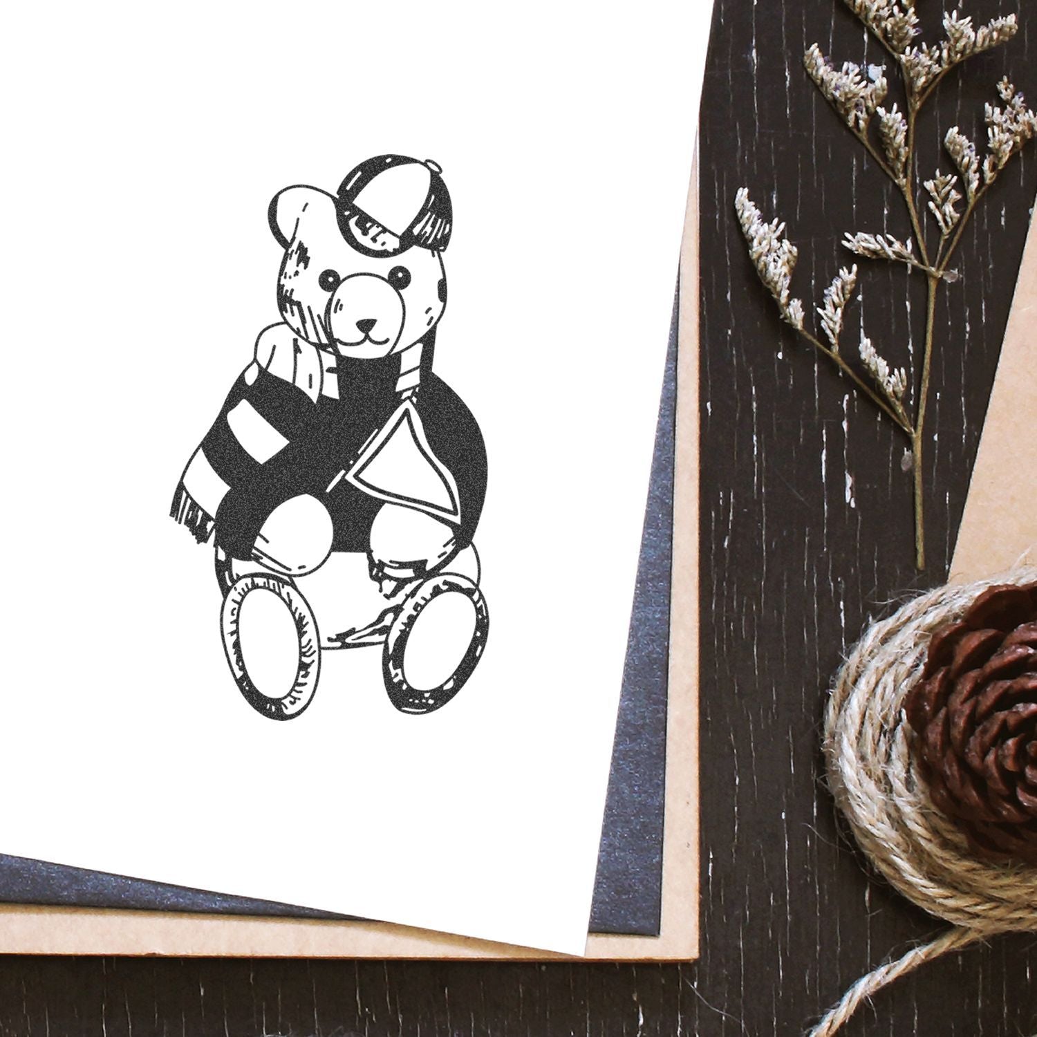Cute teddy bear illustration on white paper with rustic stationery, pinecone, twine, and dried foliage on a dark wooden background.