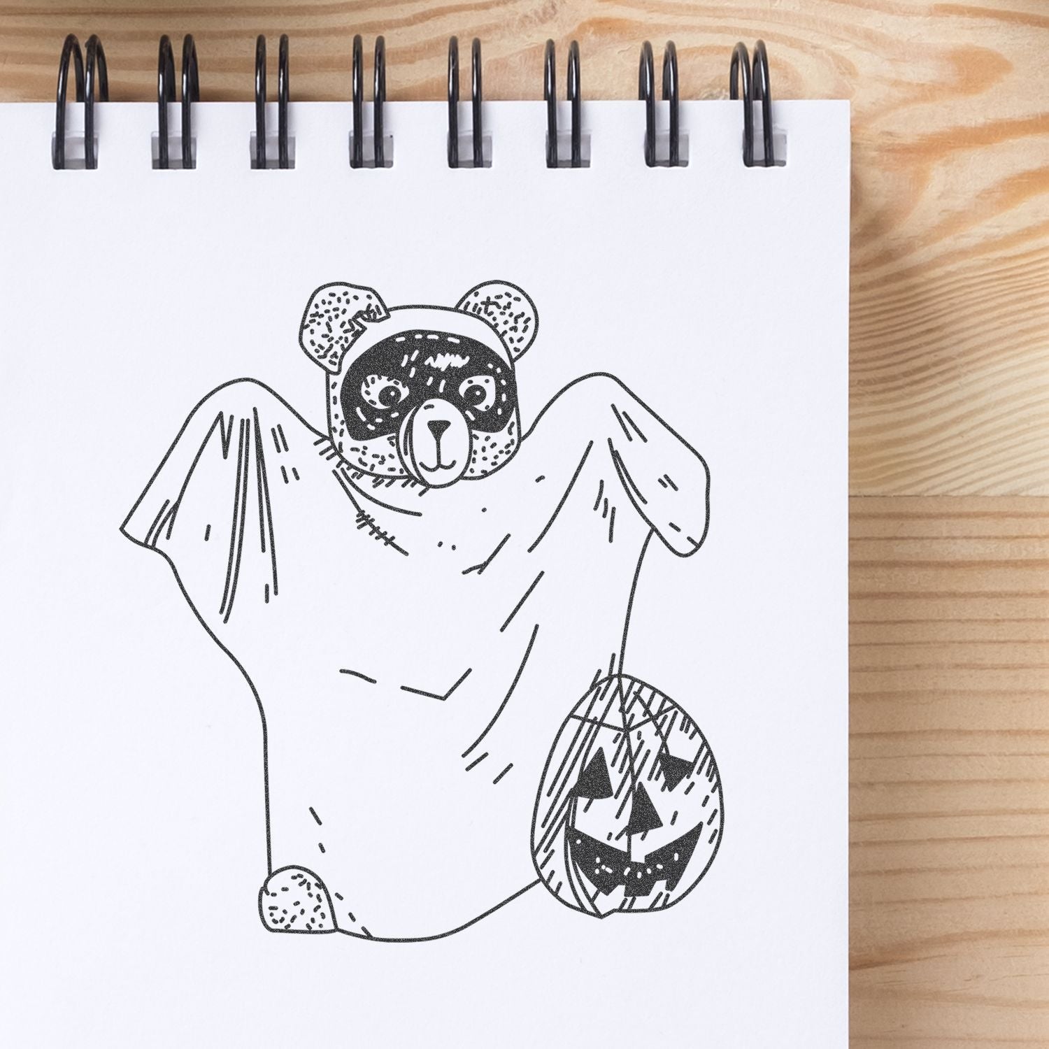 Black-and-white Halloween bear illustration in a spiral sketchbook, wearing a ghost costume and holding a jack-o-lantern pumpkin.