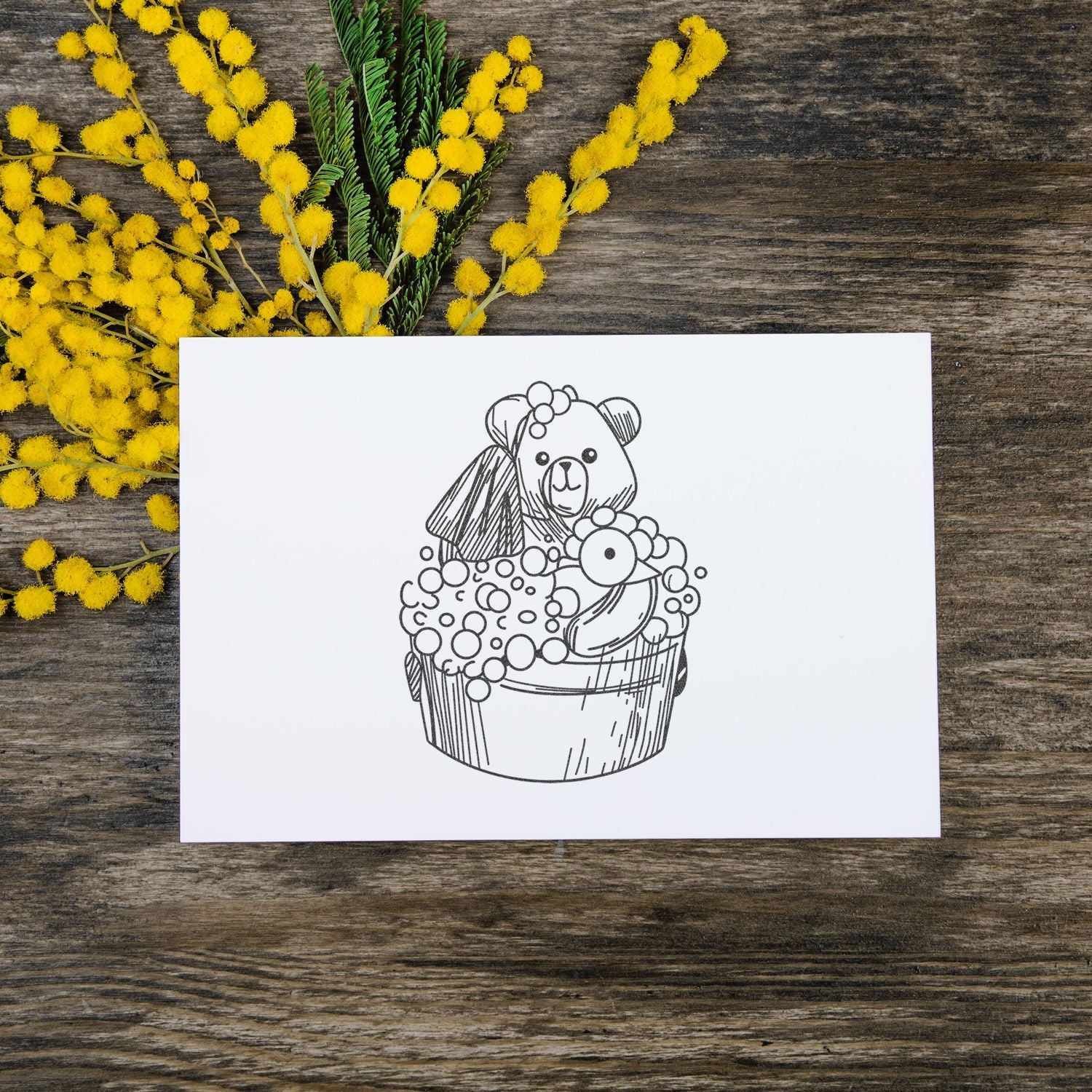Handmade greeting card with cute bear cupcake illustration on rustic wood, accented by yellow mimosa flowers.