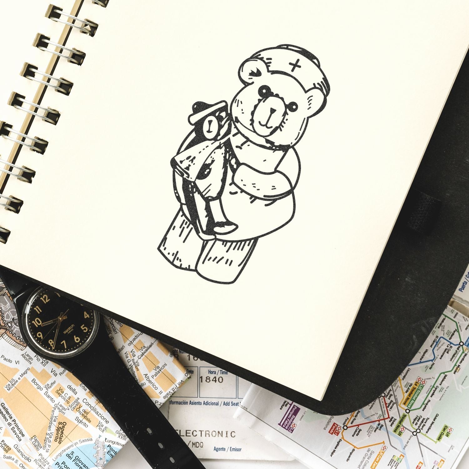 Open sketchbook with cute teddy bear illustration, surrounded by a wristwatch and travel maps on a desk.