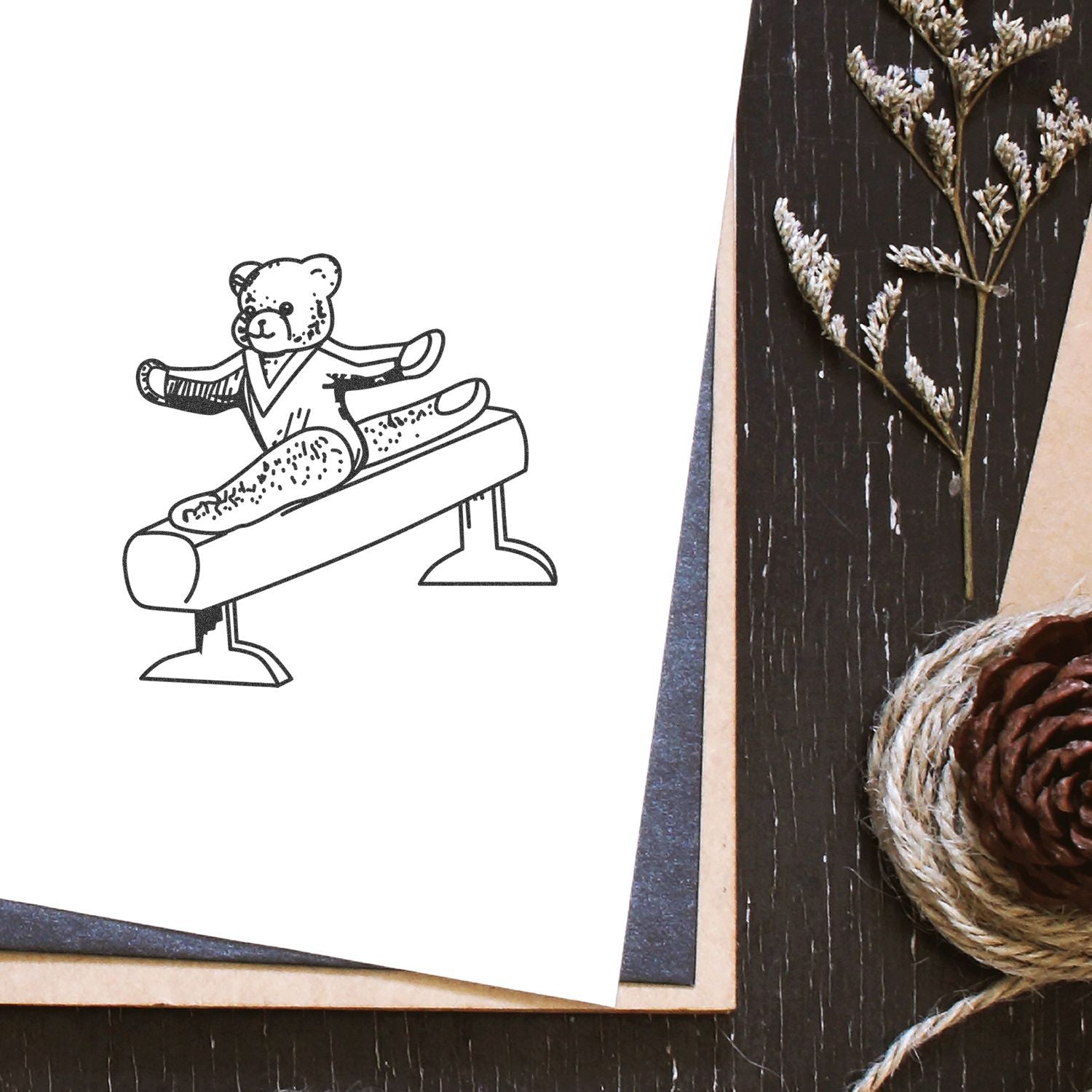 Cute teddy bear gymnastics line art on paper with rustic craft decor, dried flowers, pinecone, and twine on a wooden table.