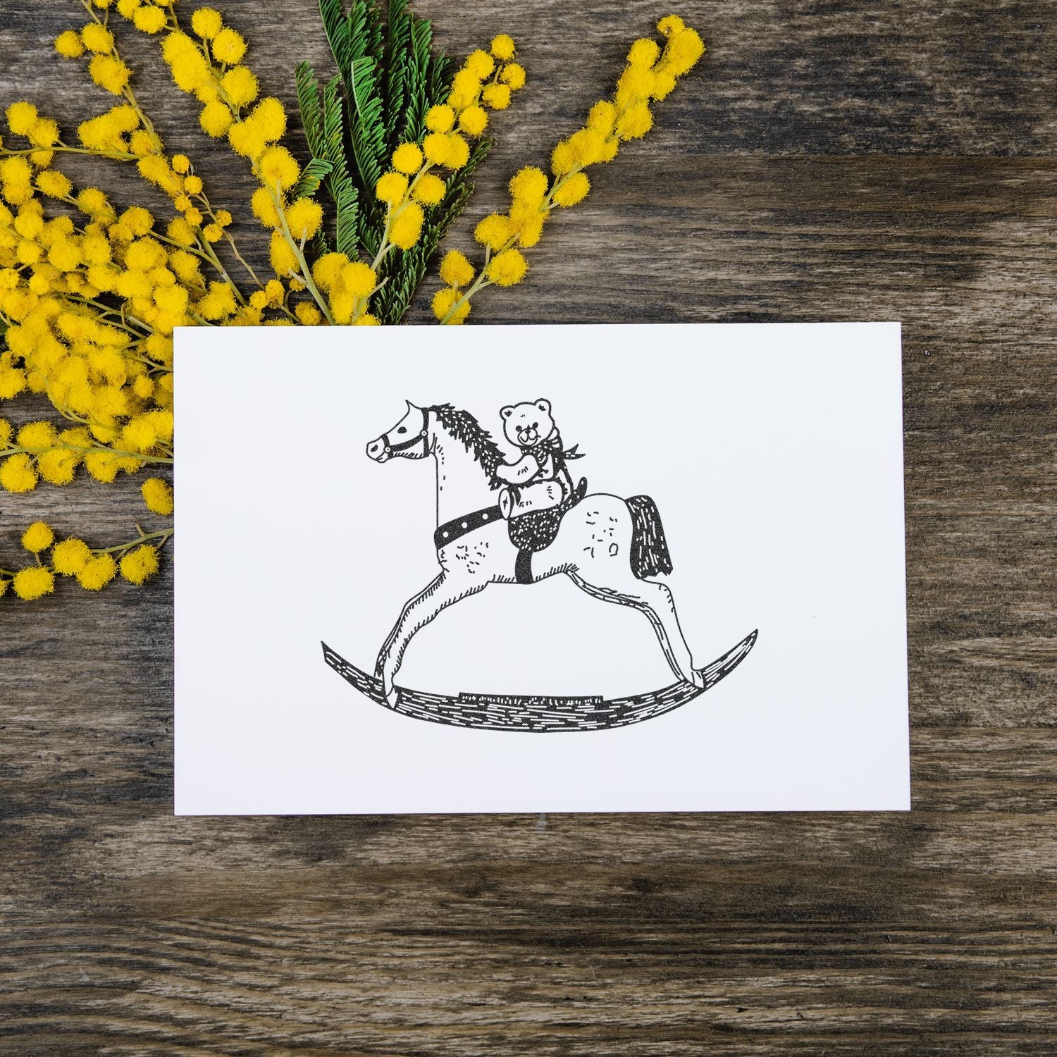 Blank greeting card with vintage rocking horse illustration and yellow mimosa flowers on rustic wood background